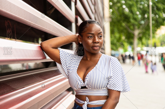 Modern young African American female in stylish striped blouse looking away while standing near railing of modern building on city street in summer day