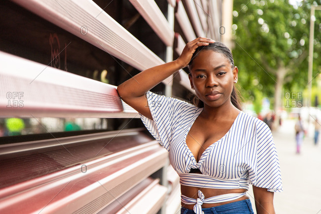 Modern young African American female in stylish striped blouse looking at camera while standing near railing of modern building on city street in summer day