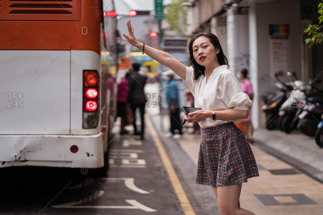 Asian female standing near road and catching cab with raised hand in Taichung city while looking forward