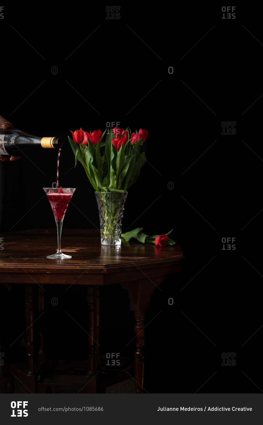 Unrecognizable person with bottle poring sparkling wine in glass placed on table with bouquet of red tulips on black background