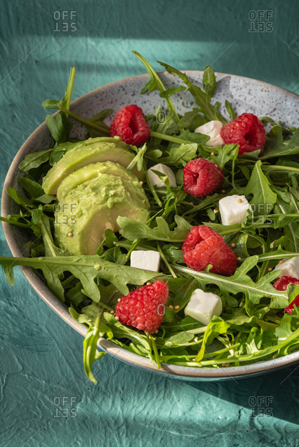 Delicious healthy salad with arugula and avocado garnished with cheese and fresh raspberries on green table