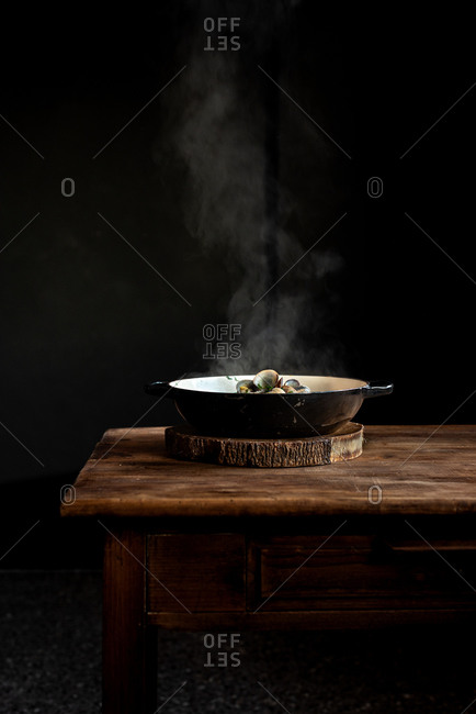 Frying pan with hot steaming freshly cooked dish placed on rustic wooden table against black background