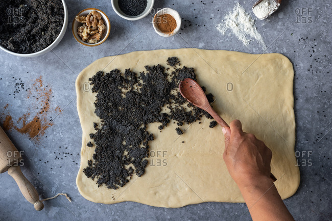 From above of crop anonymous person with wooden spoon spreading poppy seeds on rolled dough while preparing sweet pastry at table with ingredients