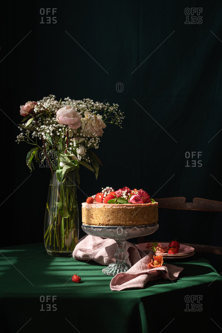 Still life composition with delicious homemade rustic cake decorated with fresh berries and flowers served on table near glass vase with delicate rose bouquet against black background