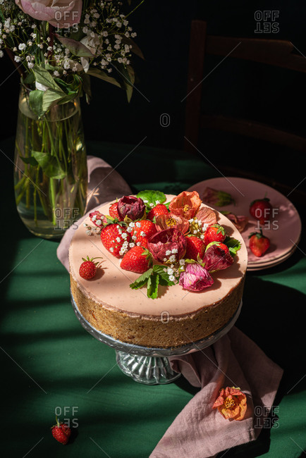 Still life composition with delicious homemade rustic cake decorated with fresh berries and flowers served on table near glass vase with delicate rose bouquet against black background