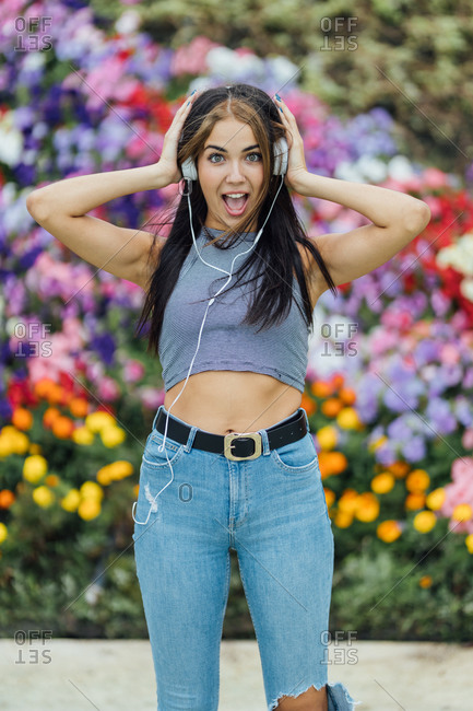 Happy amazed young female in trendy ripped jeans enjoying loud music through headphones while resting in summer garden with colorful blooming flowers
