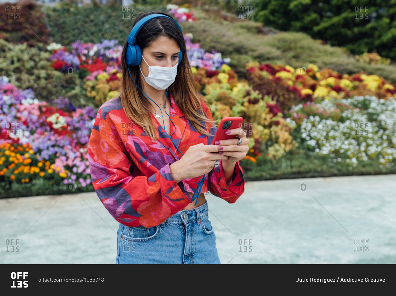 Trendy millennial female student in stylish casual outfit and medical mask with headphones communicating via mobile phone while standing in summer park with blooming flowers