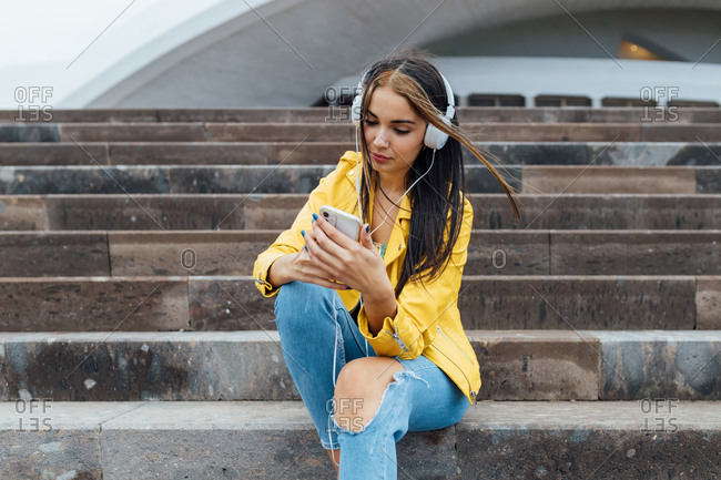 Joyful millennial woman in trendy colorful outfits and headphone sitting on stone stairs browsing smartphone while spending time together in city