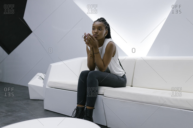 Calm African American female sitting on sofa in lobby of hotel in futuristic style while relaxing and drinking coffee