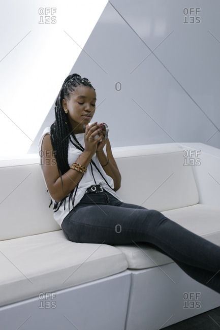 Calm African American female sitting on sofa in lobby of hotel in futuristic style while relaxing and drinking coffee with eyes closed