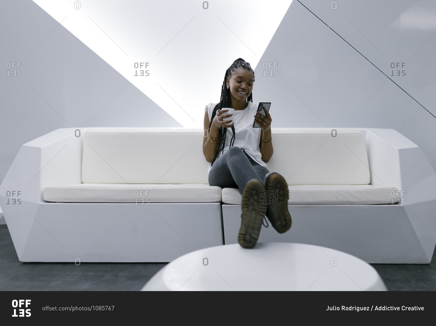 Delighted African American female sitting on sofa in room with white futuristic interior and chatting on social media on smartphone