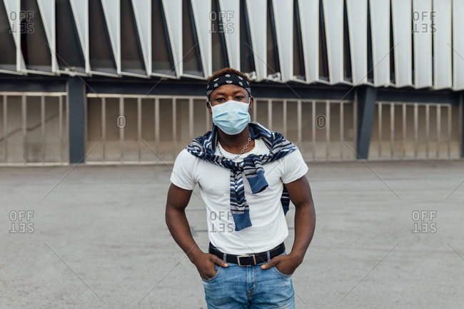 Serious African American male in casual wear with headband and protective mask looking at camera while standing against modern building on city street