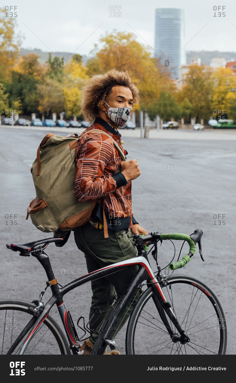 Side view of young curly haired African American man in casual clothes and protective mask with backpack standing near bicycle on paved urban square in autumn day
