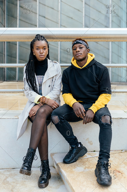 Young African American couple in trendy outfits sitting near railing looking at camera thoughtfully against contemporary urban building