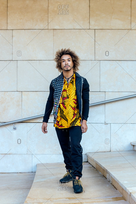 Full body of young African American male in informal trendy outfit standing on stairway against stone wall