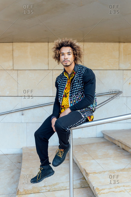 Full body of handsome curly haired bearded young male in trendy informal outfit looking at camera while sitting on metal railing near stairs of modern building