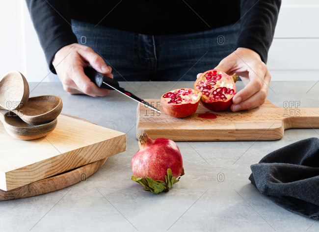 Cropped unrecognizable cook hands with knife cutting in halved fresh ripe juicy pomegranate on kitchen table in cutting board