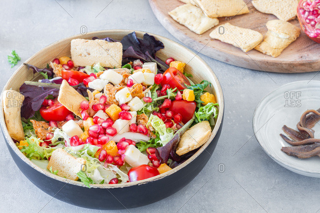 From above bowl of Caesar salad with green mix topped with cheese and tomatoes with pomegranate seeds and crackers