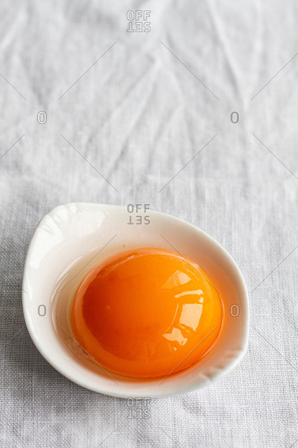 High angle of small white bowl with uncooked egg yolk placed on white tablecloth in kitchen