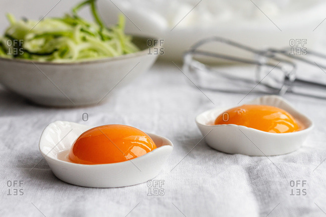Ceramic bowls with separated raw egg yolks placed on table with fresh greens and mixer during breakfast preparation