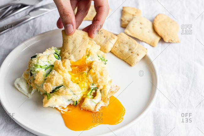 Crop anonymous person dipping cracker to yolk while eating healthy delicious homemade cloud eggs for breakfast