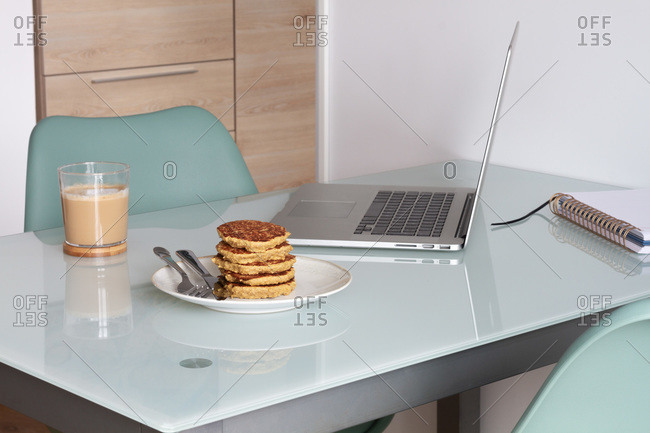 Empty table with laptop and notepad near prepare coffee and biscuits in light kitchen at home