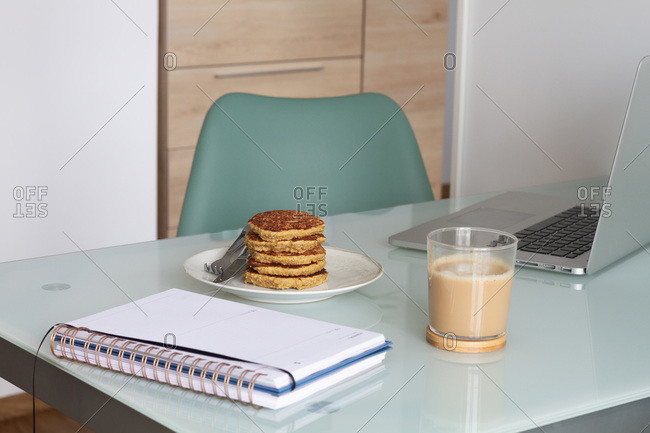 Empty table with laptop and notepad near prepare coffee and biscuits in light kitchen at home