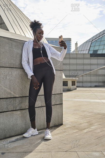 Slim African American female athlete standing on street on sunny day and messaging on mobile phone while having break during workout