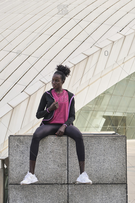 Determined African American female athlete in sportswear sitting on street and looking away