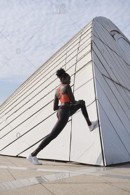 Low angle side view of flexible African American female runner warming up legs while leaning on wall and stretching body before training
