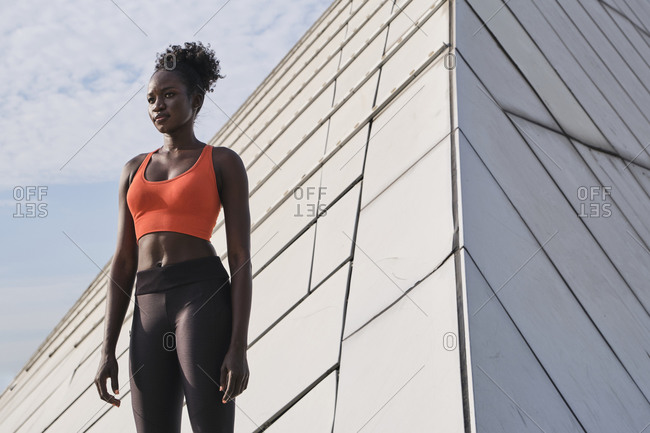 Confident African American sportswoman with slim body standing near geometric construction in city and looking away