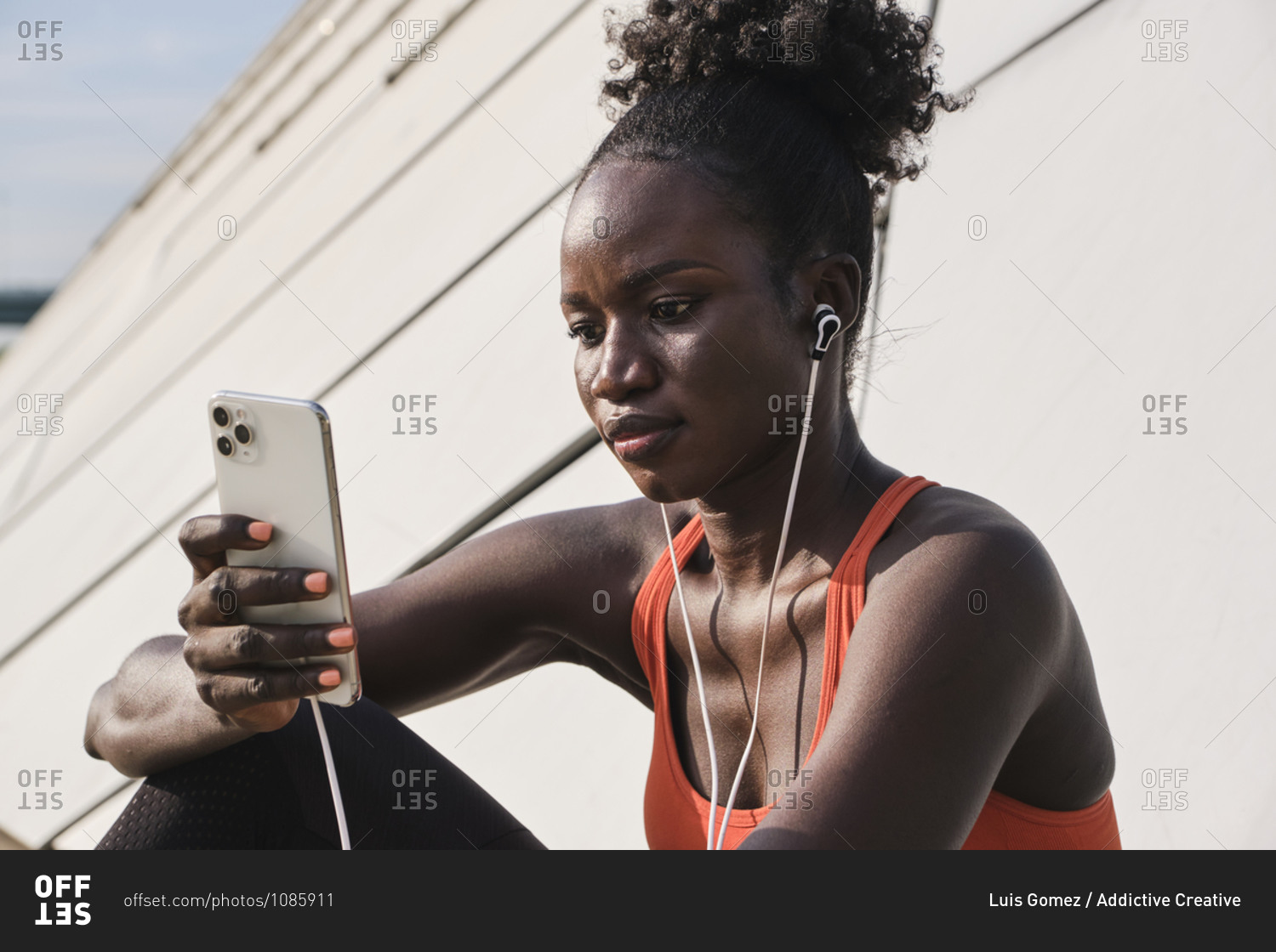 Calm African American athletic female sitting on street and listening to songs in earphones while chatting on social media via smartphone