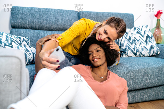 Happy young multiracial women in casual clothes cuddling and taking selfie on smartphone while spending free time together in cozy living room