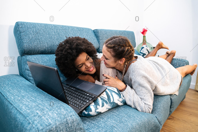 Cheerful young multiracial lesbian couple having fun while lying on sofa and browsing laptop together during weekend at home