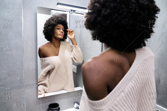 Young curly haired African American female in casual wear standing in bathroom and looking at mirror reflection during daily morning routine at home