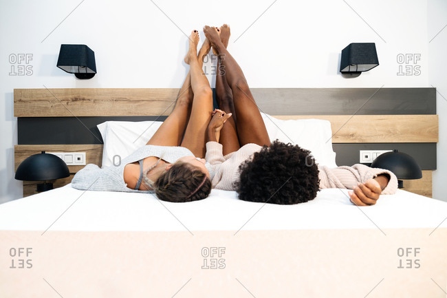 Relaxed multiracial homosexual girlfriends lying on bed with legs raised and holding hands while chilling together in bedroom at home