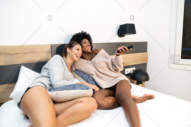 Laughing young multiracial lesbian girlfriends with remote controller watching funny TV show together while resting in bedroom