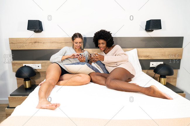 Full length of positive young multiethnic lesbian couple in casual wear browsing mobile phones while resting together on bed during free time at home