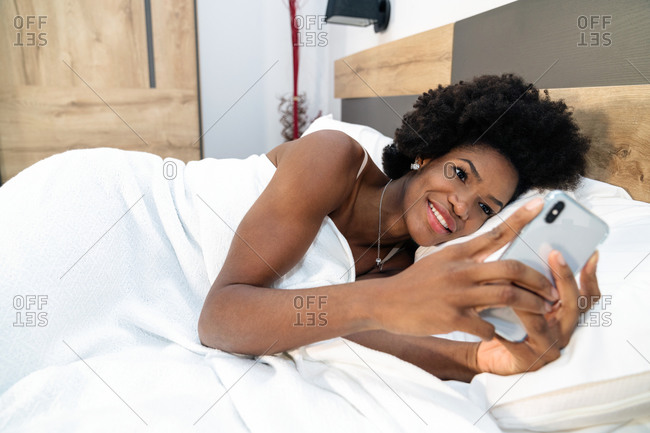 Smiling African American female lying in bed in morning and browsing mobile phone after awakening