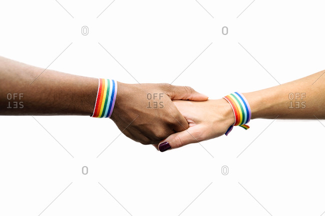 Unrecognizable crop multiracial lesbian couple with rainbow bracelets holding hands on white background