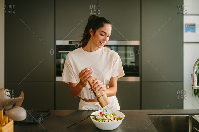 Smiling female using pepper mill and adding aromatic seasoning in bowl with homemade vegetable salad prepared for lunch