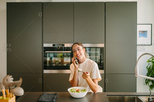 Peaceful female sitting at table in kitchen and eating tasty vegetable salad while talking on smartphone