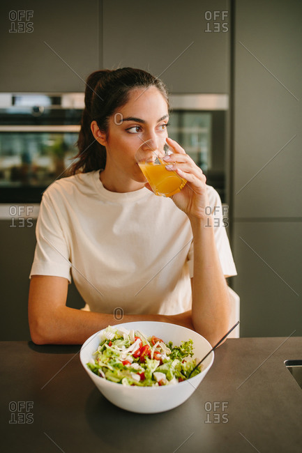 Female drinking orange juice for lunch while sitting at table with bowl of salad and looking away