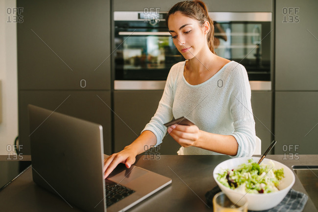 Positive young female with credit card in hand using laptop and making online order from internet shop while sitting in modern kitchen at home
