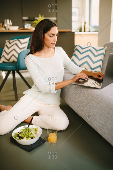Young female in casual outfit sitting on floor and working on laptop while having healthy lunch with salad and cup of juice at home