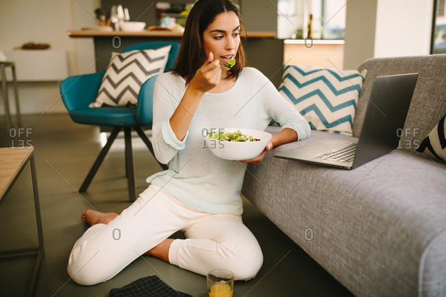 Young female in casual outfit sitting on floor and watching video on laptop while having healthy lunch with salad and cup of juice at home