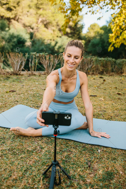 Positive young female yoga instructor setting smartphone on tripod while preparing for online yoga session in park