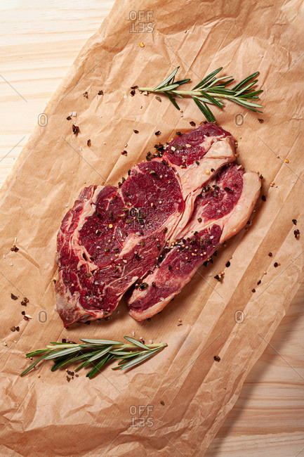 Top view of fresh uncooked juicy rib eye beef steak with fresh rosemary and pepper placed on baking paper sheet on table
