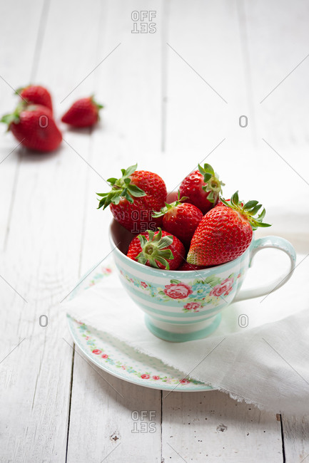 Still life with fresh ripe aromatic red strawberries in porcelain cup with saucer served for breakfast on white wooden table with tablecloth