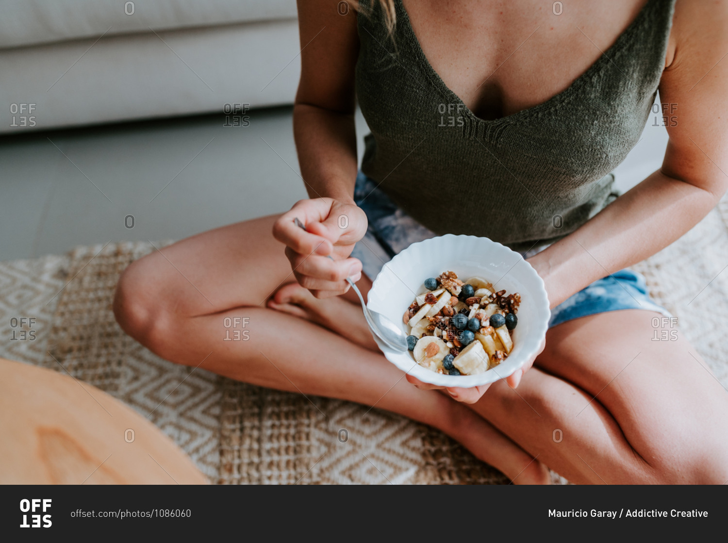 Cropped unrecognizable female with tasty bowl of healthy granola sitting on floor at home and having nourishing breakfast
